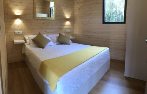 Acogedora habitación de cabaña de madera en Cabañas Compostela con cama queen, cojines oliváceos y vista al bosque en Santiago de