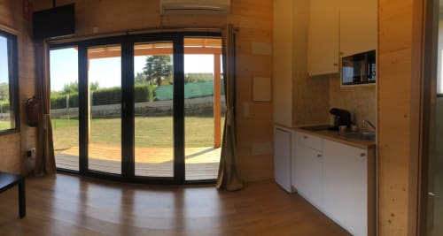 Interior de cabaña moderna con acabados en madera, zona de estar abierta, kitchenette y puertas de cristal que se abren a terraza con jardín