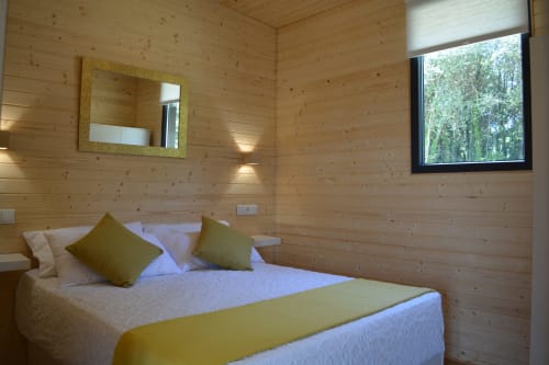 Acogedora habitación de madera en Cabañas Compostela con cama matrimonial, detalles amarillos, espejo y ventana con vista al bosque