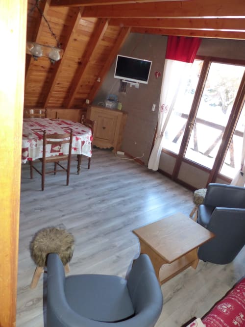 Intérieur de chalet rustique avec poutres en bois, coin salon, table à manger, TV écran plat et portes-fenêtres donnant sur balcon avec vue montagne