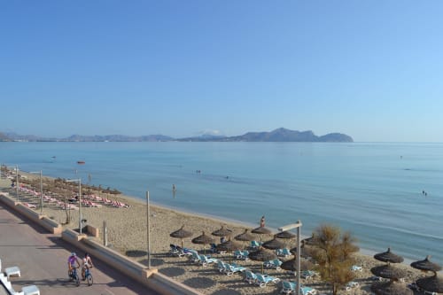 Beach at Hotel Sa Roqueta in Can Picafort with turquoise waters, umbrellas, and distant mountains under clear sky