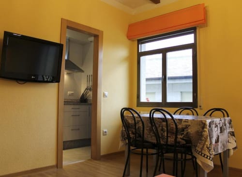 Sala de jantar amarela vibrante com cadeiras pretas, mesa, janela com cortina laranja, TV na parede e entrada para cozinha compacta