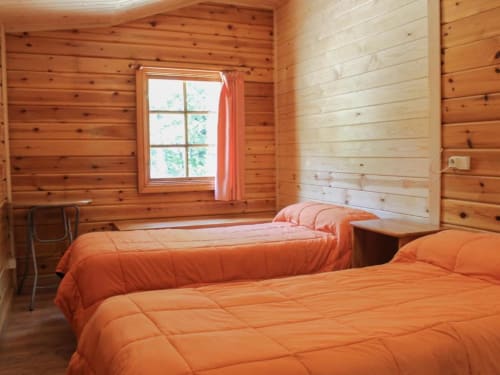 Acogedor dormitorio de cabaña de madera en Turismo en Sanabria con dos camas naranjas, ventana con cortinas rojas y luz natural