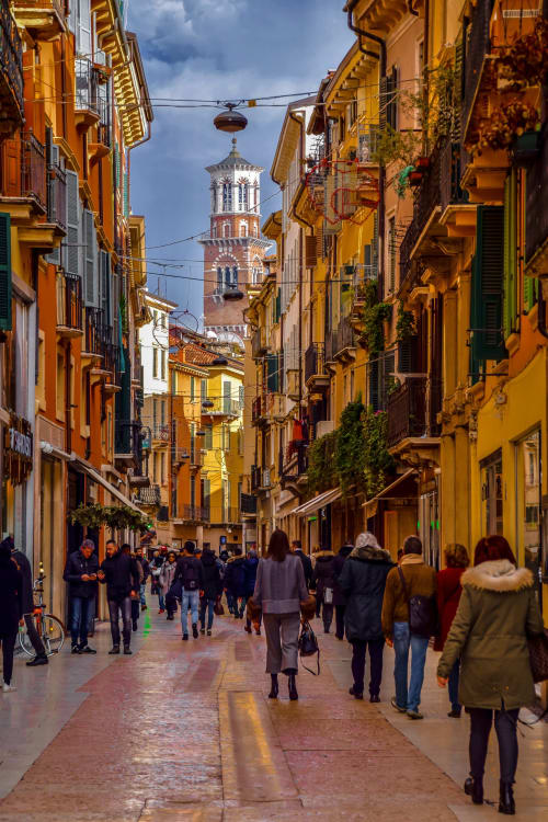 Colorful yellow buildings line a pedestrian street in Verona, Italy, with a clock tower visible in the distance and crowds of