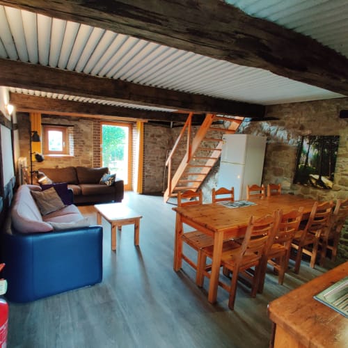 Spacious open-plan living area at La Ferme De Werpin with wooden beams, dining table, sofa, blue armchair, and stone walls