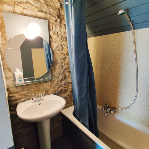 Bathroom with pedestal sink, round mirror, blue shower curtain, bathtub, and rustic stone wall at La Ferme De Werpin