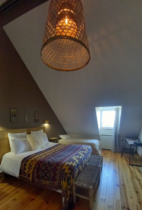 Chambre mansardée à La Maison De Fleurat avec poutres apparentes, lampe suspendue tissée, couvre-lit à motifs et fenêtre de toit