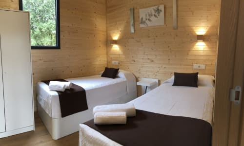 Luminosa habitación de cabaña en Cabañas Compostela con dos camas, sábanas blancas, almohadas oscuras, iluminación moderna y vistas al jardín