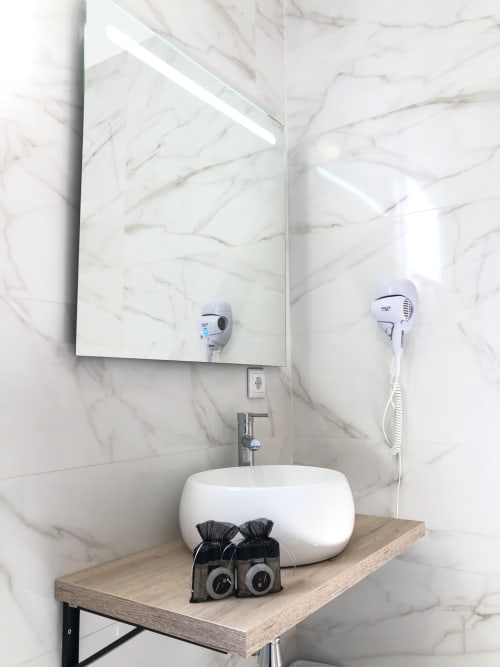 Baño moderno con lavamanos redondo blanco, paredes de mármol, espejo iluminado y secador de cabello en Cabañas Compostela