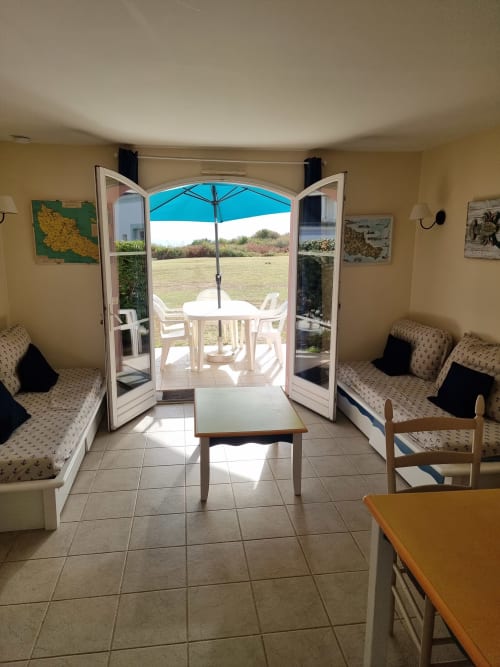 Intérieur d'appartement ensoleillé avec portes ouvertes sur la terrasse, mobilier blanc et parasol bleu surplombant Belle-Île-en-Mer