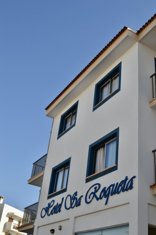 Hotel Sa Roqueta exterior facade in Can Picafort with white walls, dark blue-framed windows and balconies against clear blue