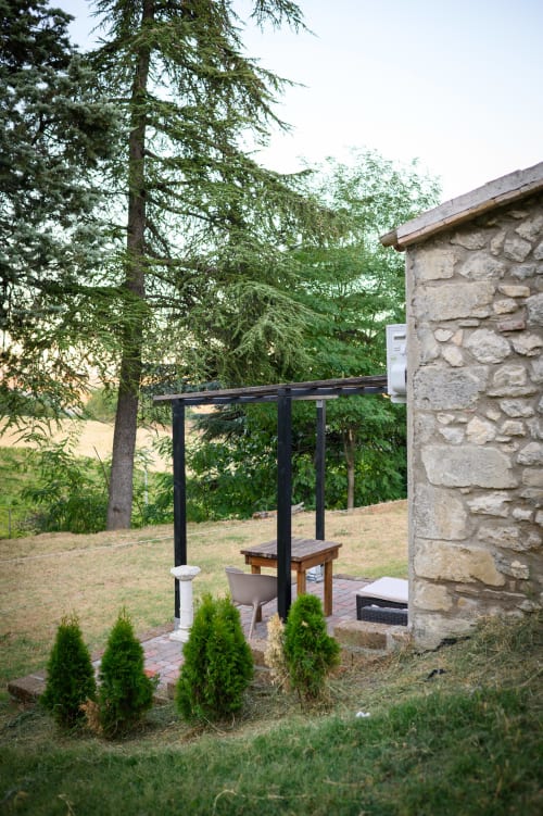 Patio in pietra con pergolato nero a Il Podere Prato, Predappio Alta, circondato da larici e aiuole curate