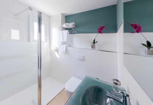 Bagno moderno con piastrelle bianche e turchesi, doccia, toilet e scaffali a parete con orchidee rosa all'Hotel Sa Roqueta