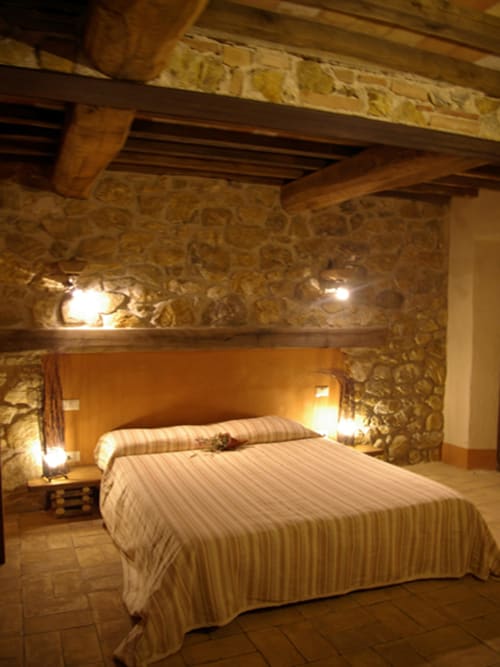 Camera da letto rustica in pietra con travi in legno, biancheria a righe e illuminazione soffusa al Casale di Poggioferro