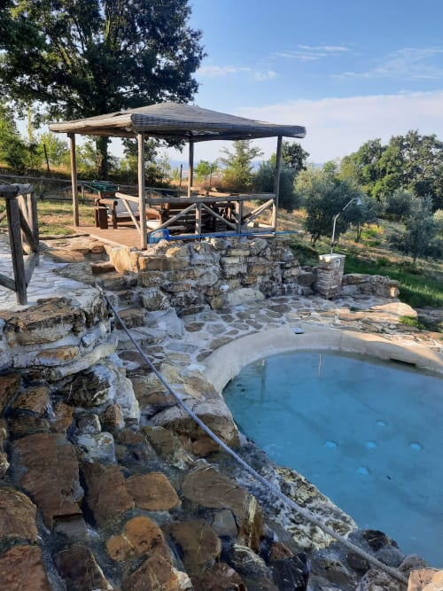 Piscina termale naturale con bordi in pietra a Casale di Poggioferro, Toscana, con pergolato in legno e zona pranzo affacciata