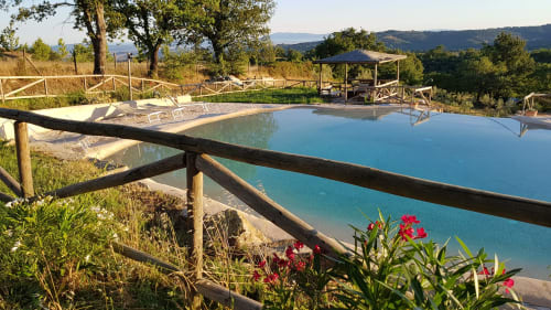 Piscina termale turchese a Casale di Poggioferro con colline toscane, ringhiere in legno e fiori rosa in primo piano