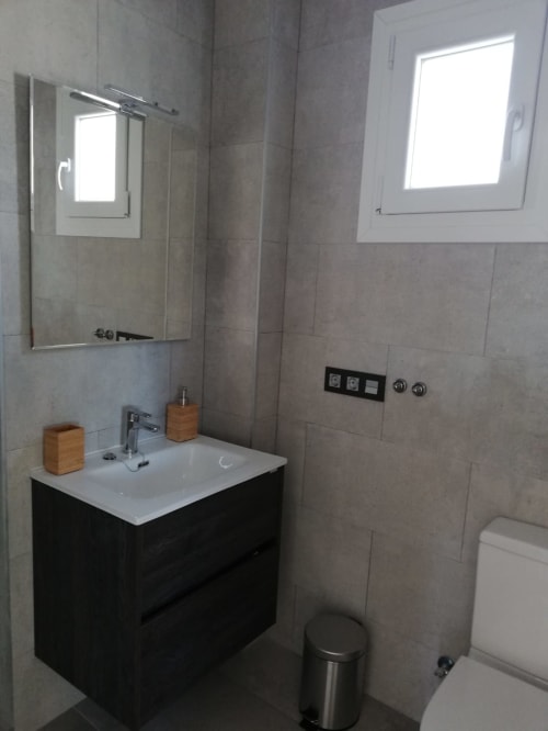 Baño moderno en El Faro De Los Genoveses con mueble negro, lavabo blanco, espejo, inodoro y paredes de azulejos grises
