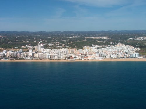 Vista aérea da vila de praia Armação de Pêra com litoral arenoso, oceano azul e apartamentos brancos