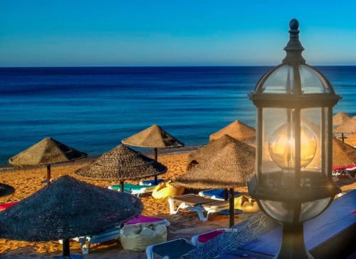 Residências de Armação ao entardecer com guarda-sóis de palha, barcos e lanterna iluminada com vista para águas azuis tranquilas.