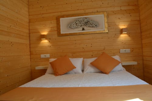 Acogedor dormitorio de cabaña de madera en Cabañas Compostela con cama matrimonial, almohadas naranjas y cuadros de árboles