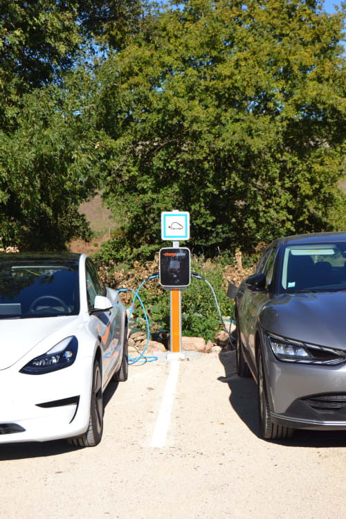 Electric car charging station at Logis De La Bénétière in Treize-Vents with two vehicles parked nearby and green trees in