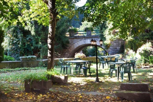 Zone de détente extérieure à la Résidence Soansa du Gérardmer à Soultzeren avec tables, chaises et arbres près d'un pont de pierre.