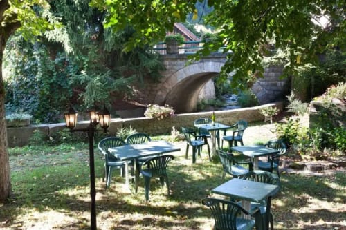 Zone de restauration extérieure à la Résidence Soansa du Gerardmer à Soultzeren avec tables, chaises et pont de pierre sur la rivière