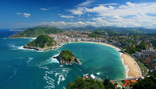 Vista aérea de bahía turquesa con playa dorada, acantilados verdes y pueblito costero entre montañas vascas