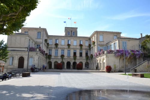 Grand courtyard of Le Pavillon M in Grignan with elegant stone buildings, purple flowers, and fountains