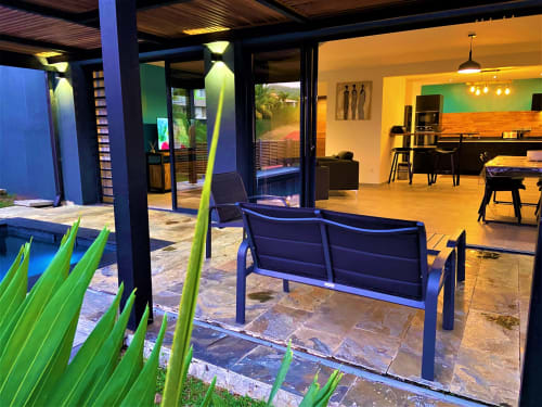 Patio de la villa Passion Cannelle avec piscine, banquette bleue, plantes vertes et espace de vie intérieur aux Hébergements Passion