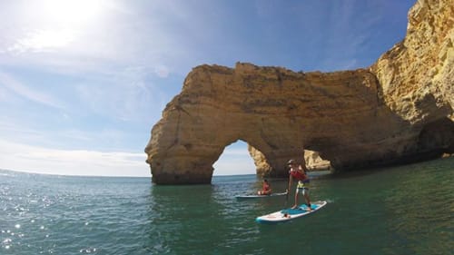Stand-up paddleboarders near natural rock arch in turquoise waters at Armação de Pêra, Portugal