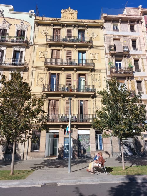 Barcelona apartment building exterior with classical architecture, tree-lined patio area, and person sitting in courtyard at