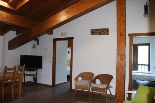 Amplio interior de apartamento en Casa Lafuente con vigas de madera, sillas de mimbre, TV y acceso a balcón con vistas a la montaña
