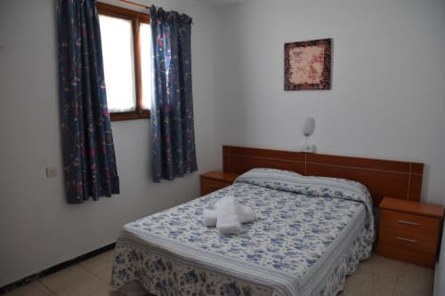 Acogedora habitación en la pensión Eva de Puerto de Mogán con cortinas estampadas azules, cama de madera y arte floral en las paredes