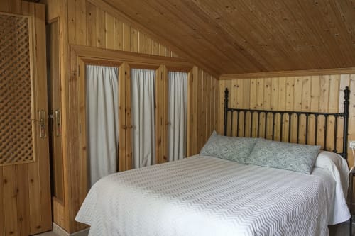 Dormitorio rústico con paneles de madera, cama de estructura metálica negra y tres ventanas con cortinas color crema en Casa Rural Las Cuevas De