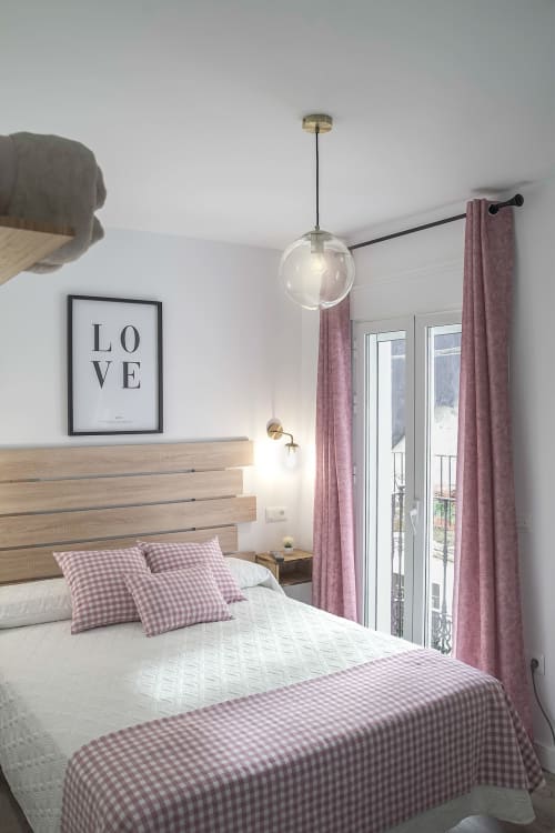 Luminoso dormitorio en Casa Rural Las Cuevas De Setenil con cama de madera, almohadas a cuadros rosas, lámpara colgante y balcón