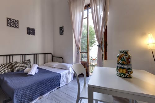 Bright Florence apartment bedroom with blue bedding, wrought iron bed, balcony doors, desk, and Tuscan pottery decor
