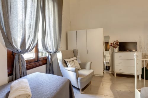 Bright Florence apartment bedroom with cream furnishings, window with grey curtains, comfortable armchair, and TV at Sleep