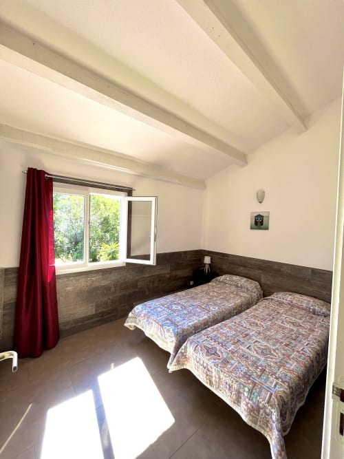 Chambre lumineuse de la villa Le Maquis Plage avec deux lits simples, boiseries sombres, rideaux rouges et vue sur le jardin