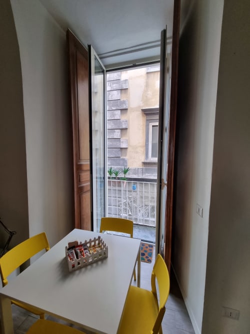 Sunny dining area in Naples suite with yellow chairs, white table, and city balcony view of historic buildings