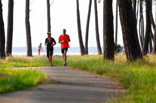Deux joggeurs courant sur un sentier bordé d'arbres près de la plage de Biscarrosse avec l'eau visible en arrière-plan
