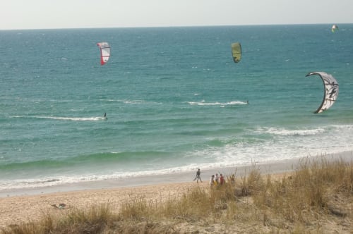 Des kitesurfeurs profitent de cerfs-volants colorés sur les eaux turquoise de la plage de Biscarrosse, avec des oyats visibles.