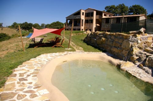 Piscina termale naturale di Casale di Poggioferro con terrazza in pietra, vela parasole rossa e villa rustica nella campagna toscana