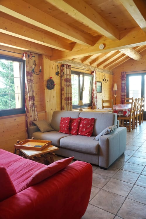 Salon douillet du chalet en bois avec canapé gris, coussins rouges et poutres rustiques à La Louvière, Fontcouverte