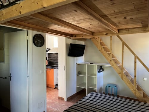 Intérieur mini villa Le Maquis Plage avec poutres en bois, escalier vers mezzanine, TV, étagères et cuisine ouverte