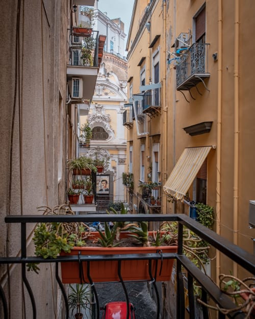 Narrow Naples alley with colorful buildings, potted plants on balcony railing, and ornate church facade - Museo19 apartment