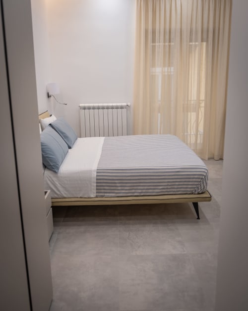 Modern bedroom at Museo19 apartment in Naples with striped bedding, radiator, and sheer curtains overlooking the city.