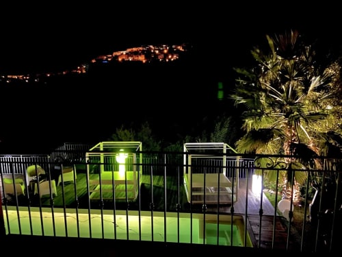 Terrasse de la piscine illuminée de la Villa Colina avec palmiers et lumières du village en montagne la nuit, Mandelieu la Napoule