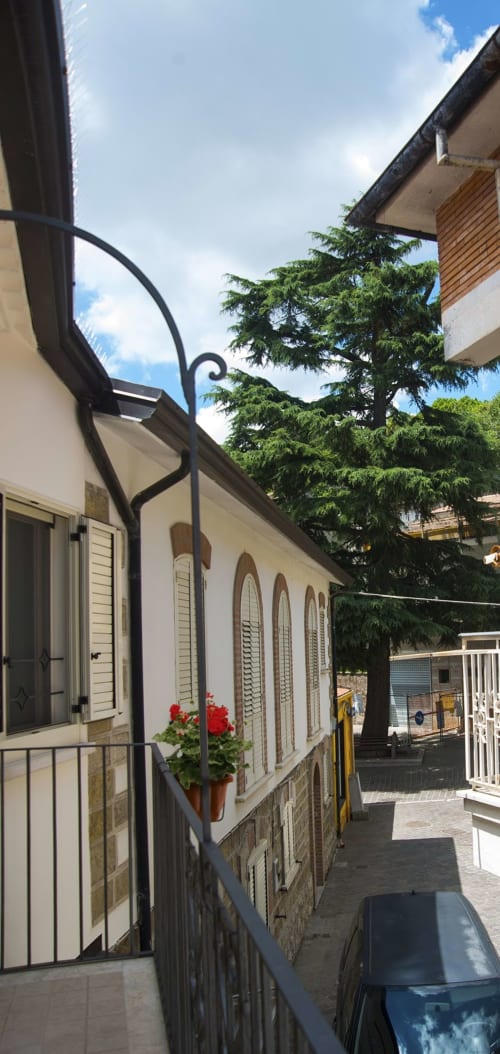 Corte di B&B La Pigna a Filiano con edifici in pietra bianca, tenda nera, fiori rossi e alberi verdi sotto cielo azzurro