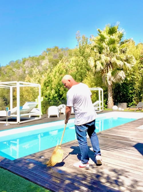 Homme balayant la terrasse de la piscine à Villa Colina, un gîte à Mandelieu-la-Napoule avec piscine turquoise et palmiers